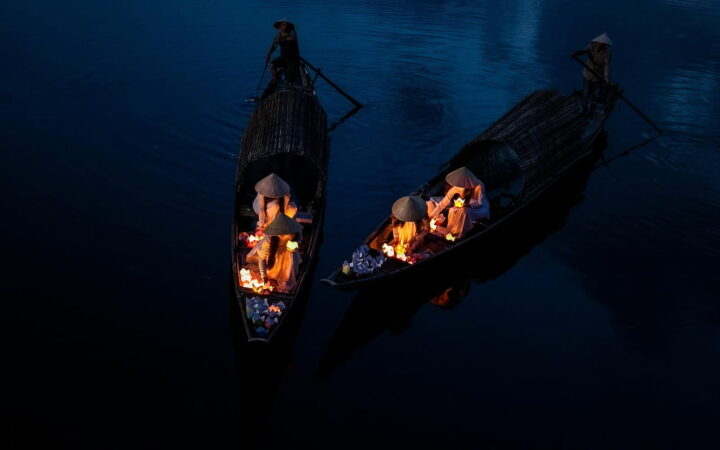 Deux barques traditionnelles naviguant à la nuit tombée lors d'une aventure au Vietnam, avec des femmes en chapeaux coniques manipulant des lanternes lumineuses sur l'eau sombre.